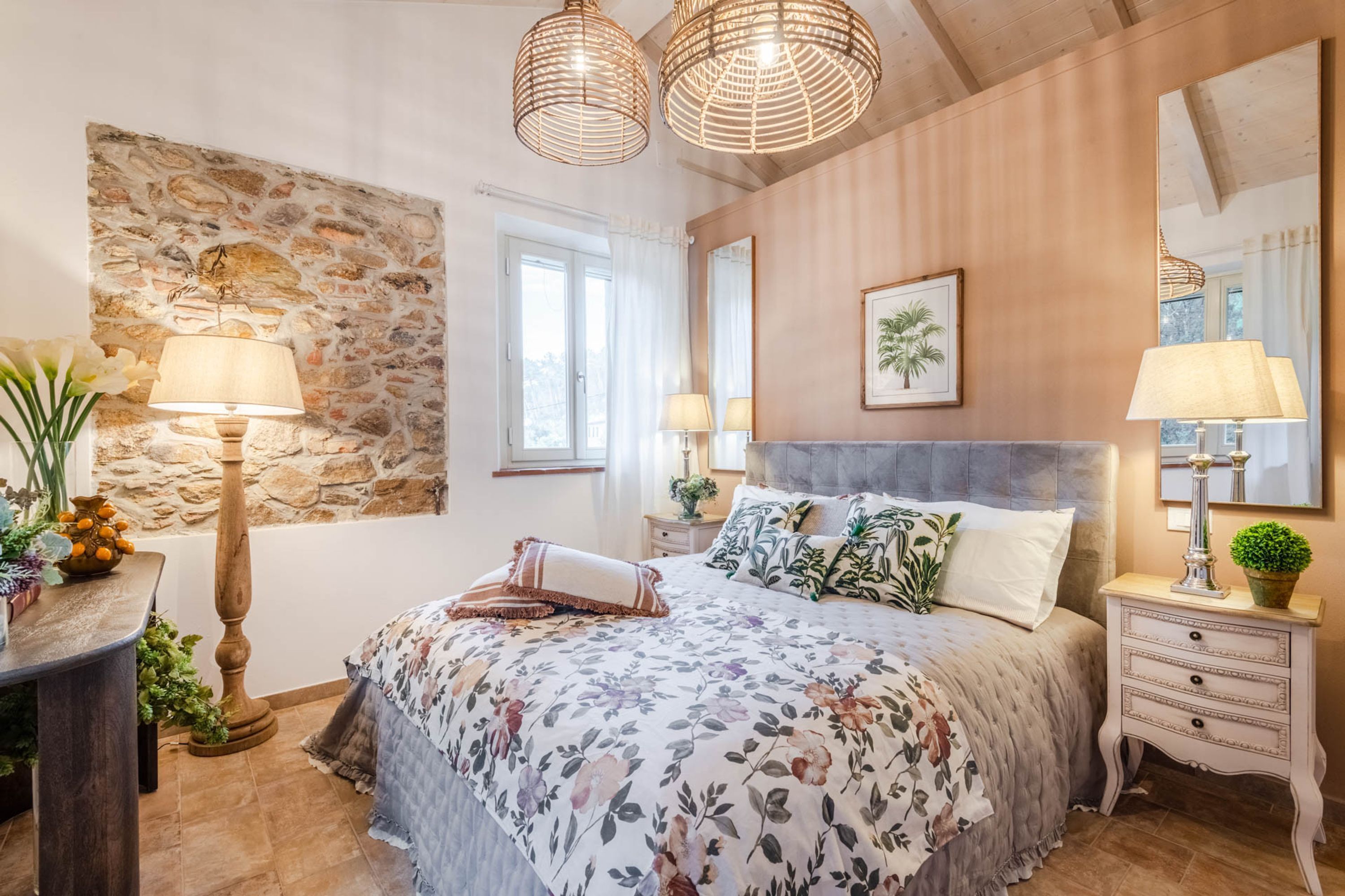 A bedroom with a stone wall, a bed with floral bedding, two bedside lamps, and a large mirror.