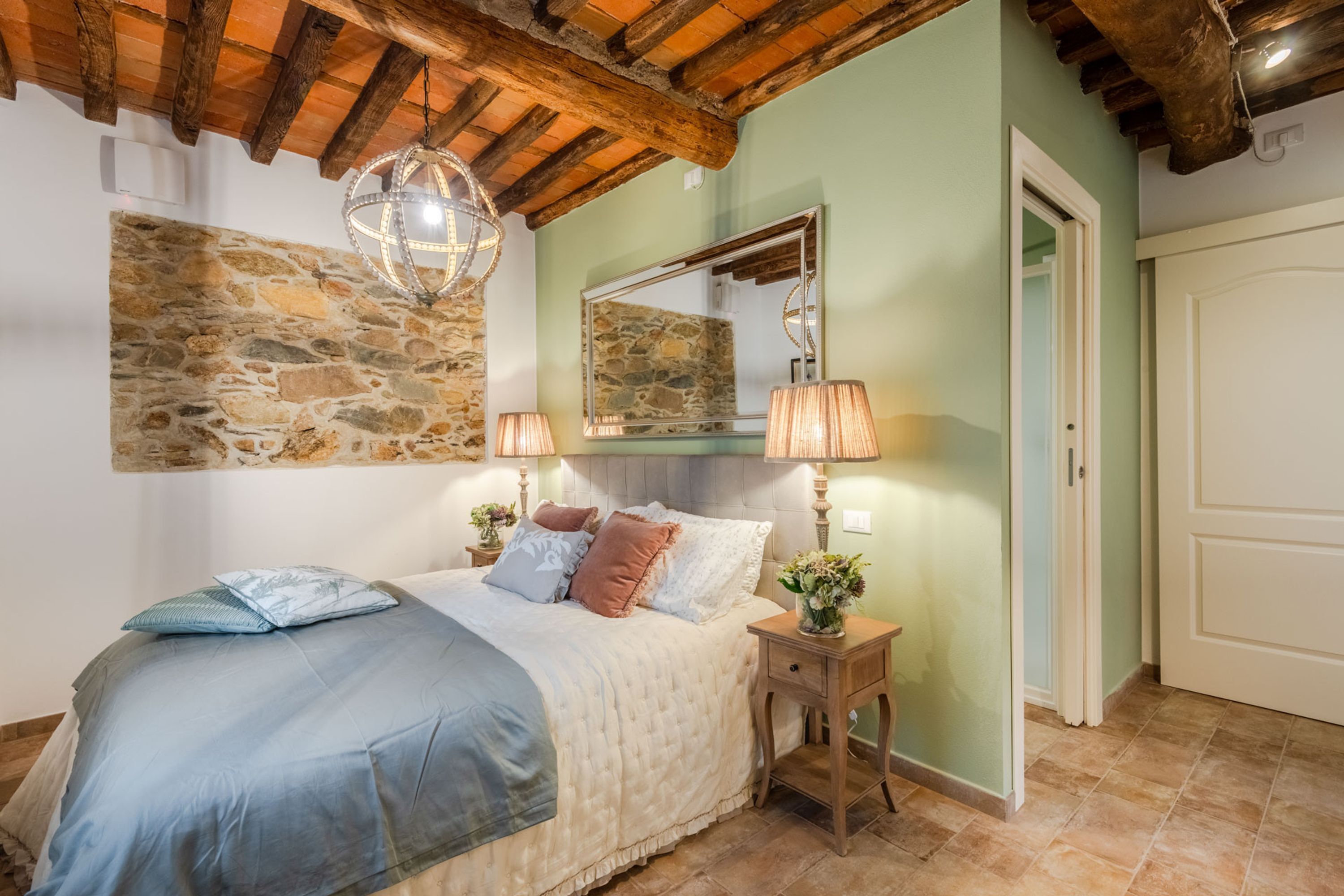 A bedroom with a stone wall, a wooden ceiling, a bed with a blue and white bedspread, two bedside lamps, and a mirror above the bed.