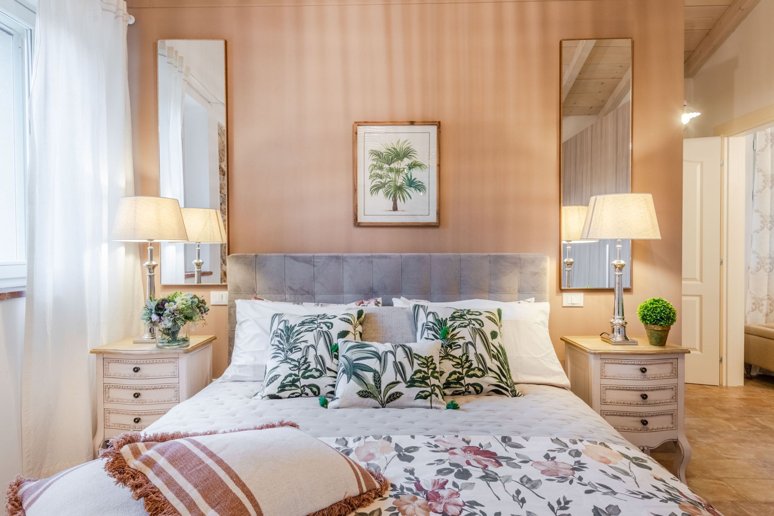 Een slaapkamer met een groot bed, bloemenbedrukking, twee nachtkastjes met lampen, een plantenprint in een lijst aan de muur en een spiegel.