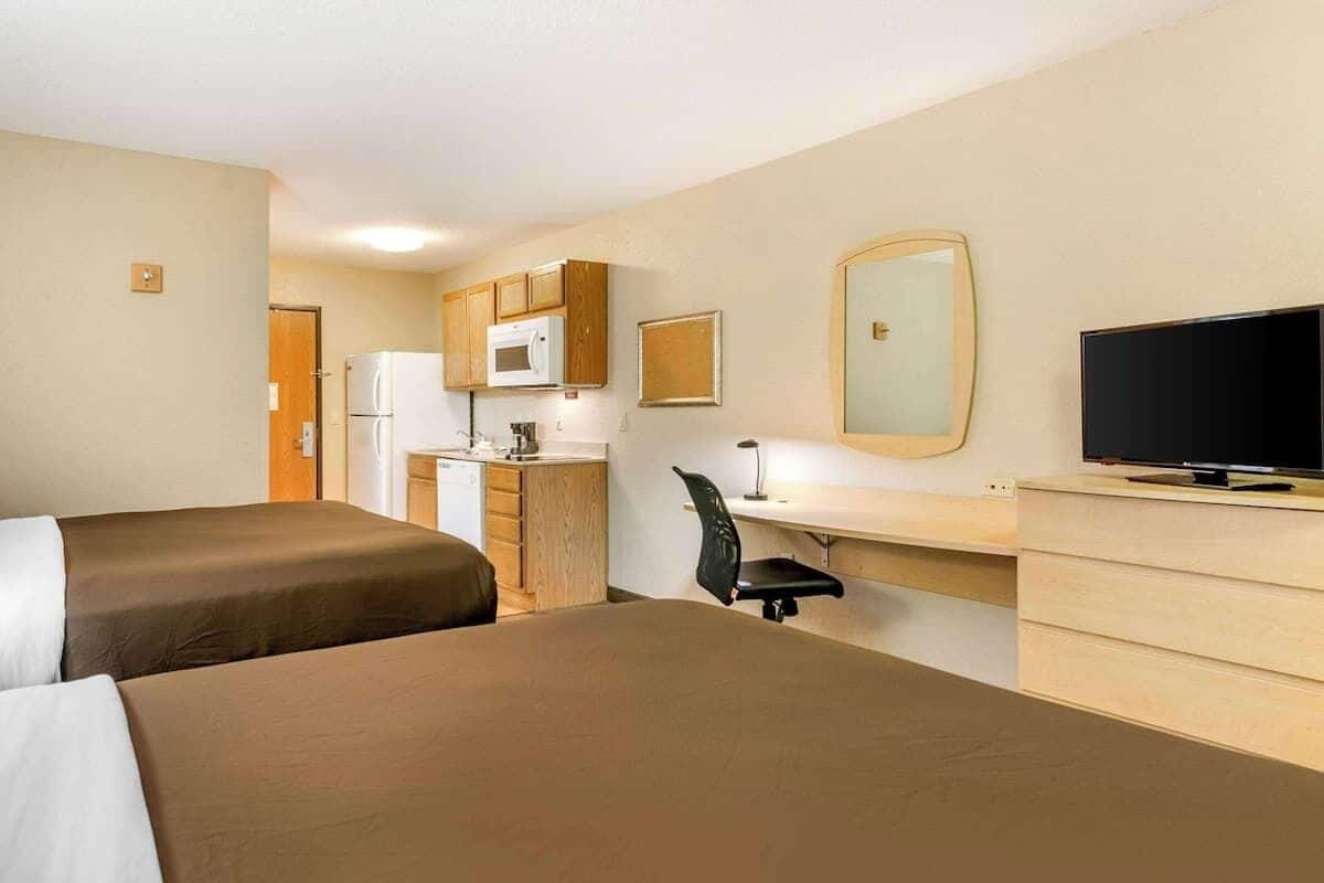 Habitación de hotel con dos camas, una kitchenette, un escritorio con silla, un televisor y un espejo.