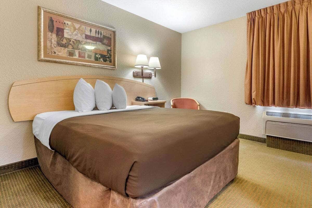 Habitación de hotel con una cama grande, cabecero, mesita de noche con lámpara, una silla y ventana con cortinas.