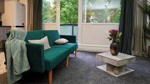 A green sofa with a blanket and pillows, a small table with a vase and cactus, and a window with curtains. - 1br, Balcony, Cityview, Office, Ws98 (London)