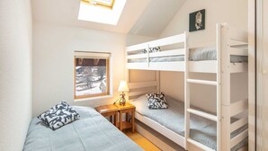 A dorm-style room with bunk beds, a small bedside table with a lamp, a window with a view of a snowy landscape, and a skylight above the beds.