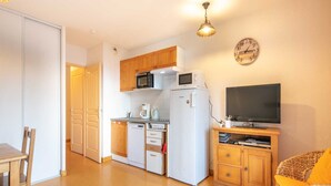 A compact kitchen with a microwave, toaster, and refrigerator, a television on a wooden stand, and a wicker chair with a yellow cushion.