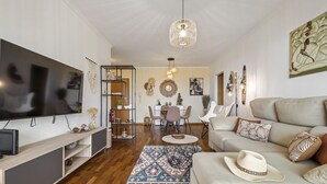A modern living room with a flat-screen TV, a sofa, a dining area, and a chandelier. - Aura Serena by Madeira Sun Travel (Santa Cruz)