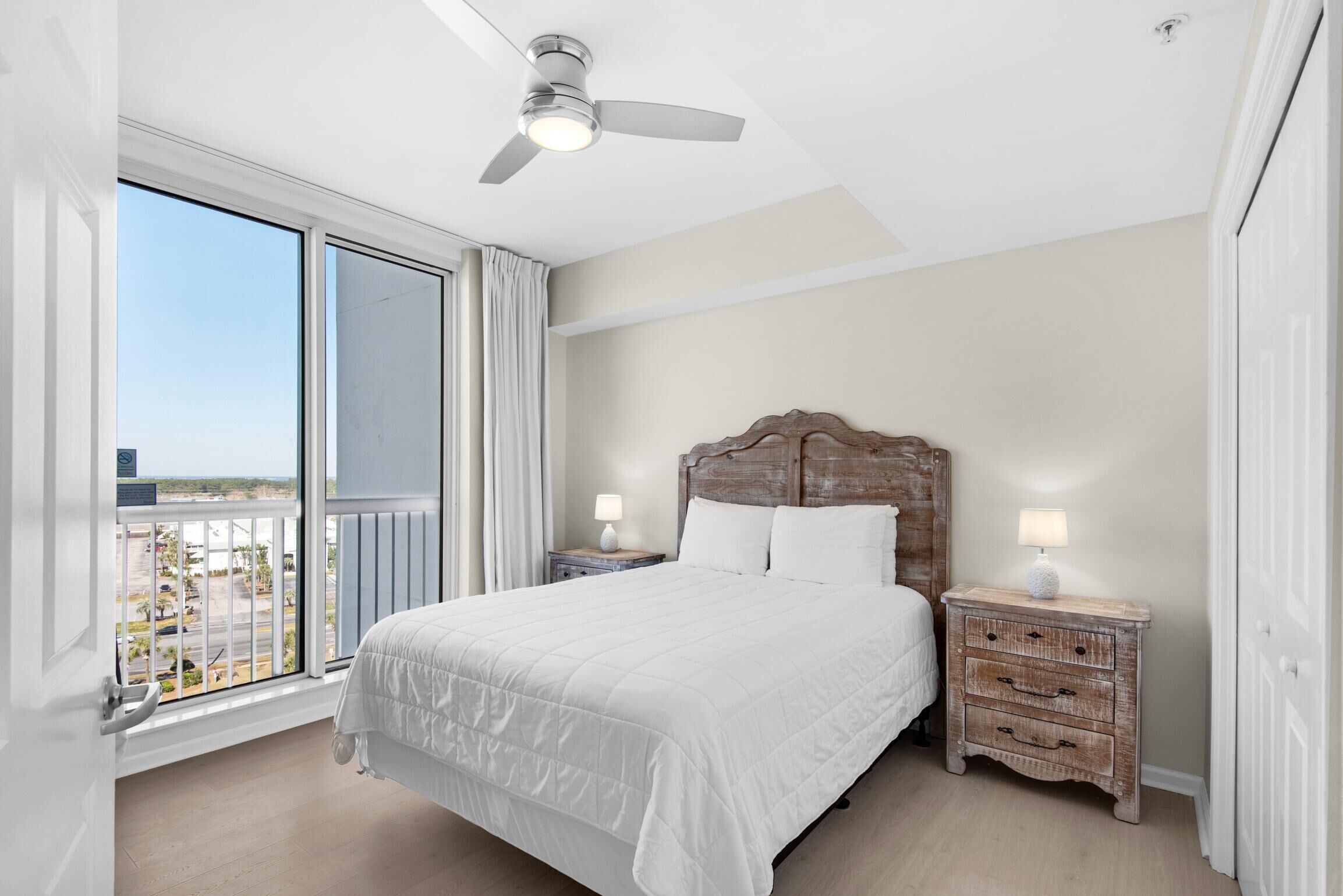 Habitación de hotel con una cama grande, cabecera de madera, dos mesitas de noche con lámparas, ventilador de techo y un balcón con vistas.