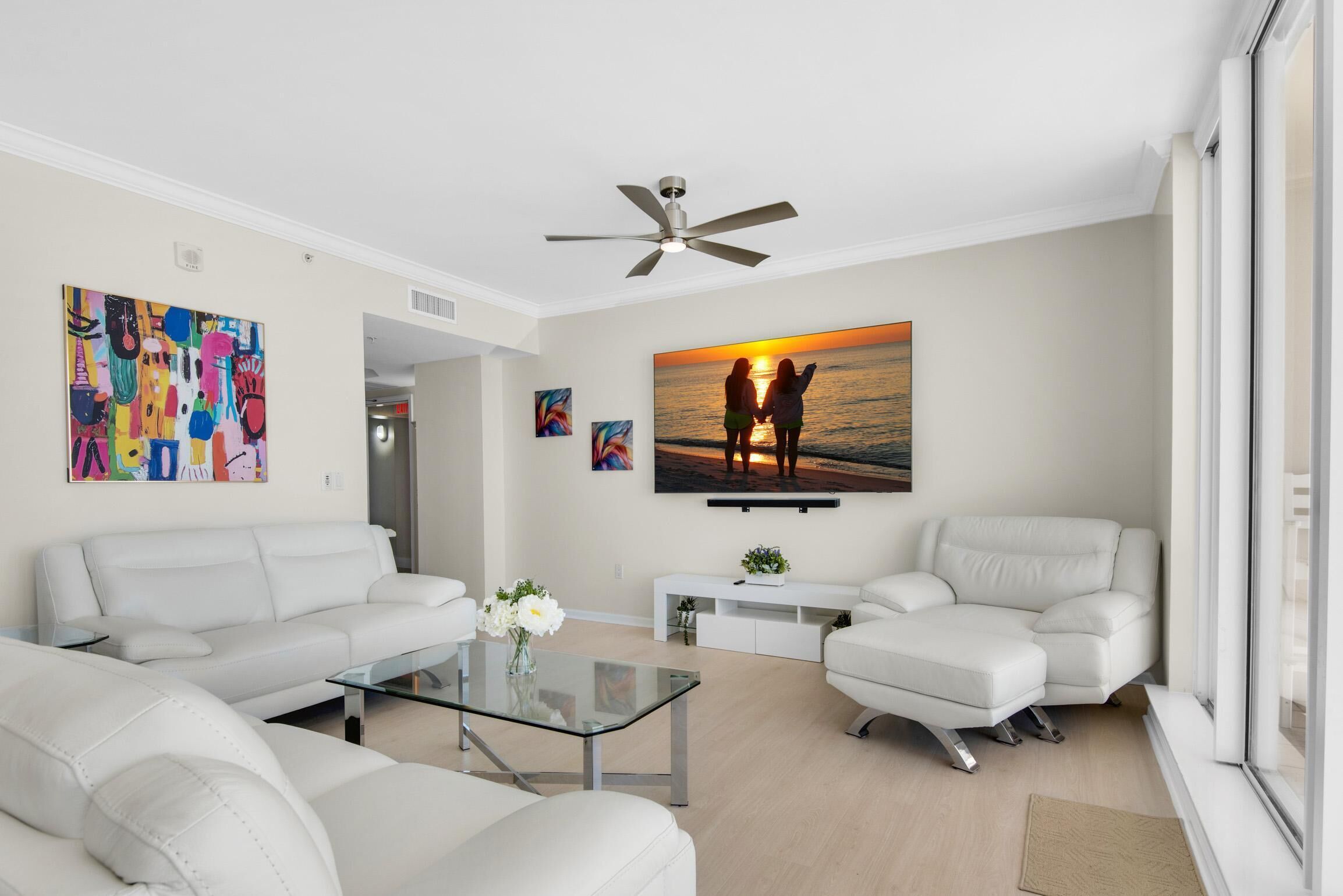 A modern living room with white sofas, a glass coffee table, and a ceiling fan.
