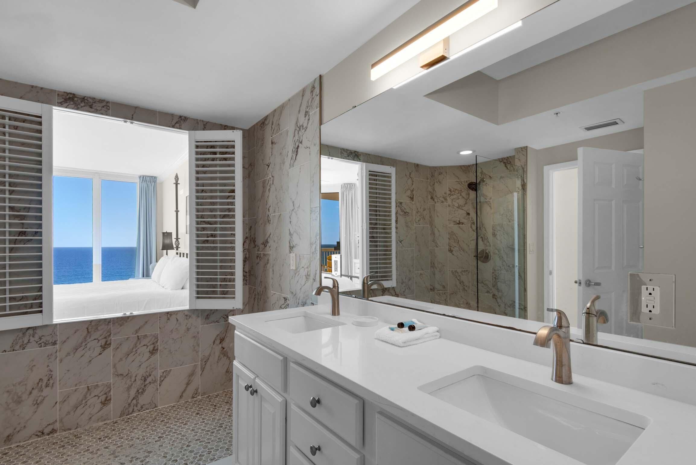 Een moderne badkamer met een grote spiegel, twee wastafels en uitzicht op zee door een raam met luiken.