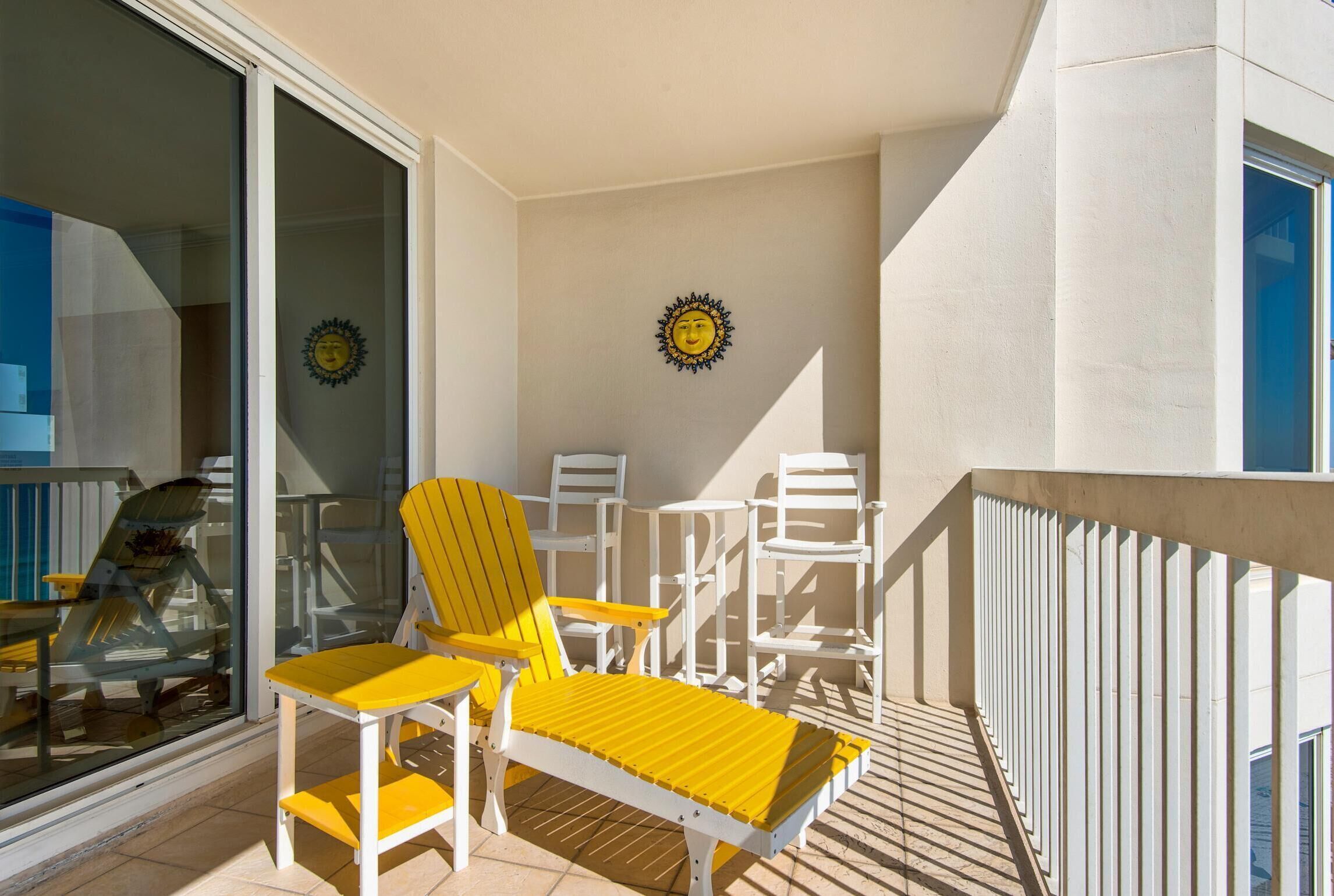 A balcony with yellow lounge chairs, white tables, and a glass door leading to a pool area.