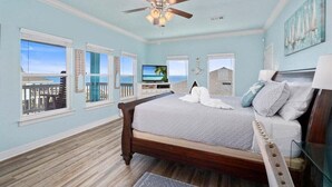 A bedroom with a bed, a ceiling fan, a TV, and a view of the ocean through large windows. - 2nd Row Crystal Beach Home | Steps Away | Sleeps 18 (Crystal Beach)