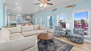 A spacious living room with a beige sectional sofa, a round coffee table, and patterned armchairs. A ceiling fan and a fish decoration are visible on the walls. - 2nd Row Crystal Beach Home | Steps Away | Sleeps 18 (Crystal Beach)