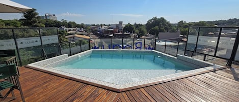 A rooftop pool with a wooden deck, surrounded by glass railings and a clear view of the cityscape.