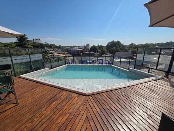 A rooftop pool with a wooden deck, surrounded by glass railings and a clear view of the cityscape.