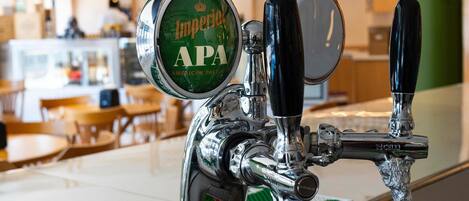Zona de bar con una grifo etiquetado como 'Imperial APA' y una pared verde con estantes llenos de botellas.
