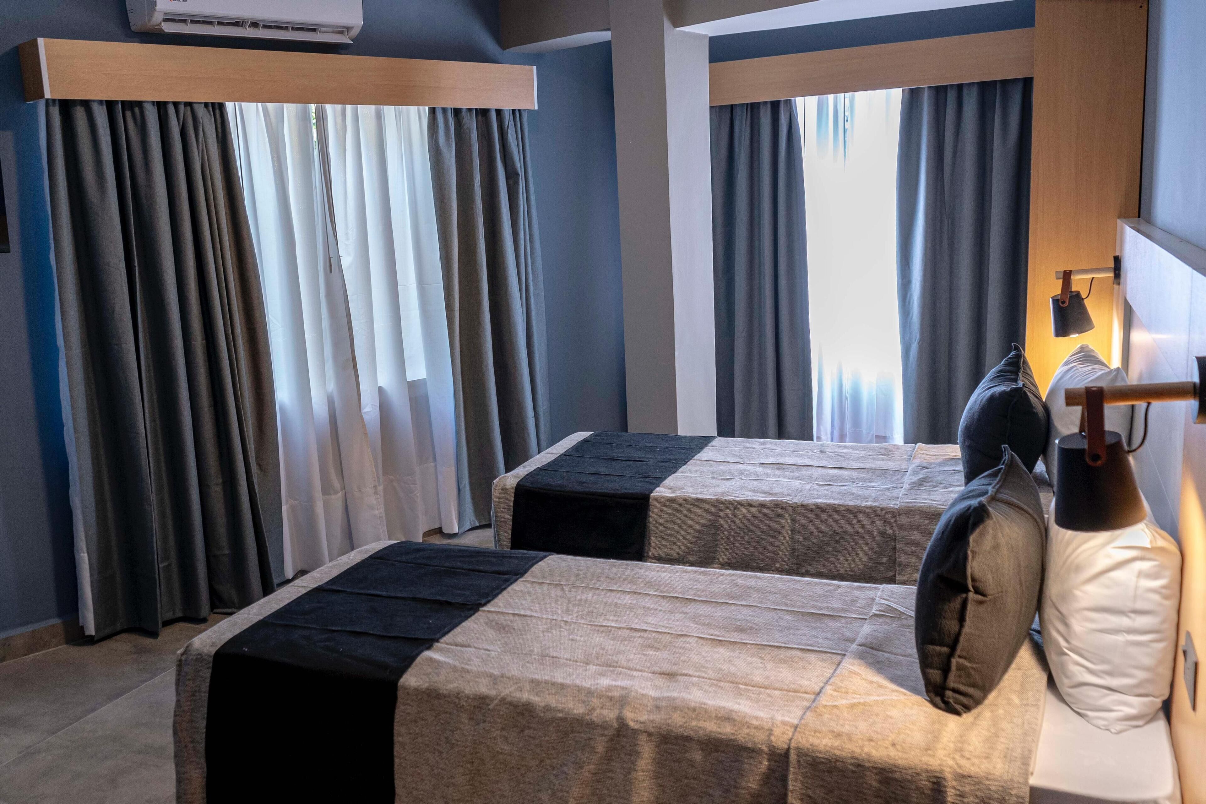 A hotel room with two beds, each with a black and beige bedspread, a bedside lamp, and a window with curtains.