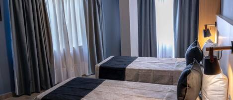 A hotel room with two beds, each with a black and beige bedspread, a bedside lamp, and a window with curtains.
