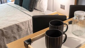 Una habitación de hotel con una cama bien hecha, una mesita de noche con dos tazas negras y un recipiente de vidrio, y una lámpara de pared.