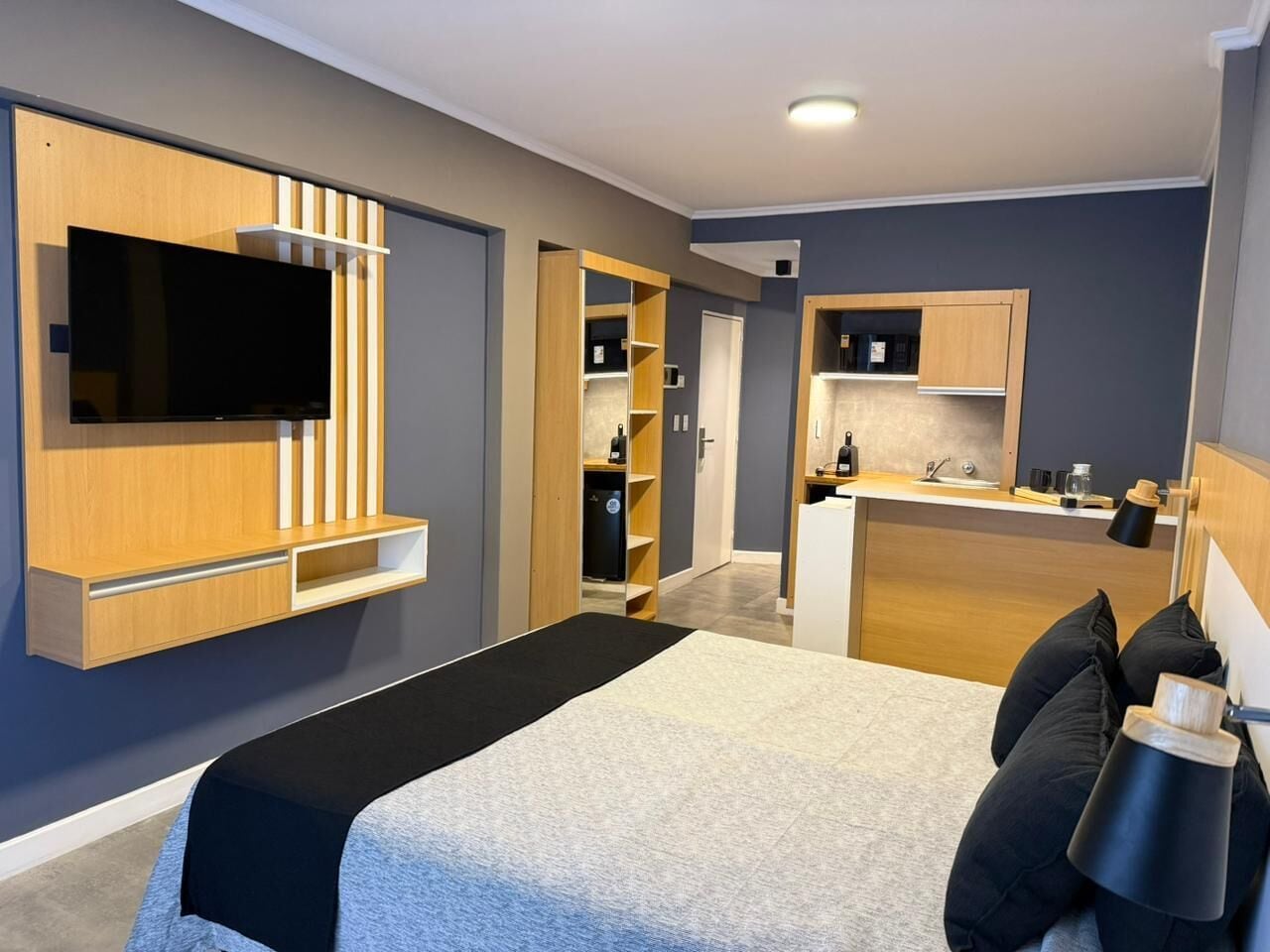 A modern hotel room with a bed, a TV mounted on the wall, a kitchenette, and a walk-in closet.