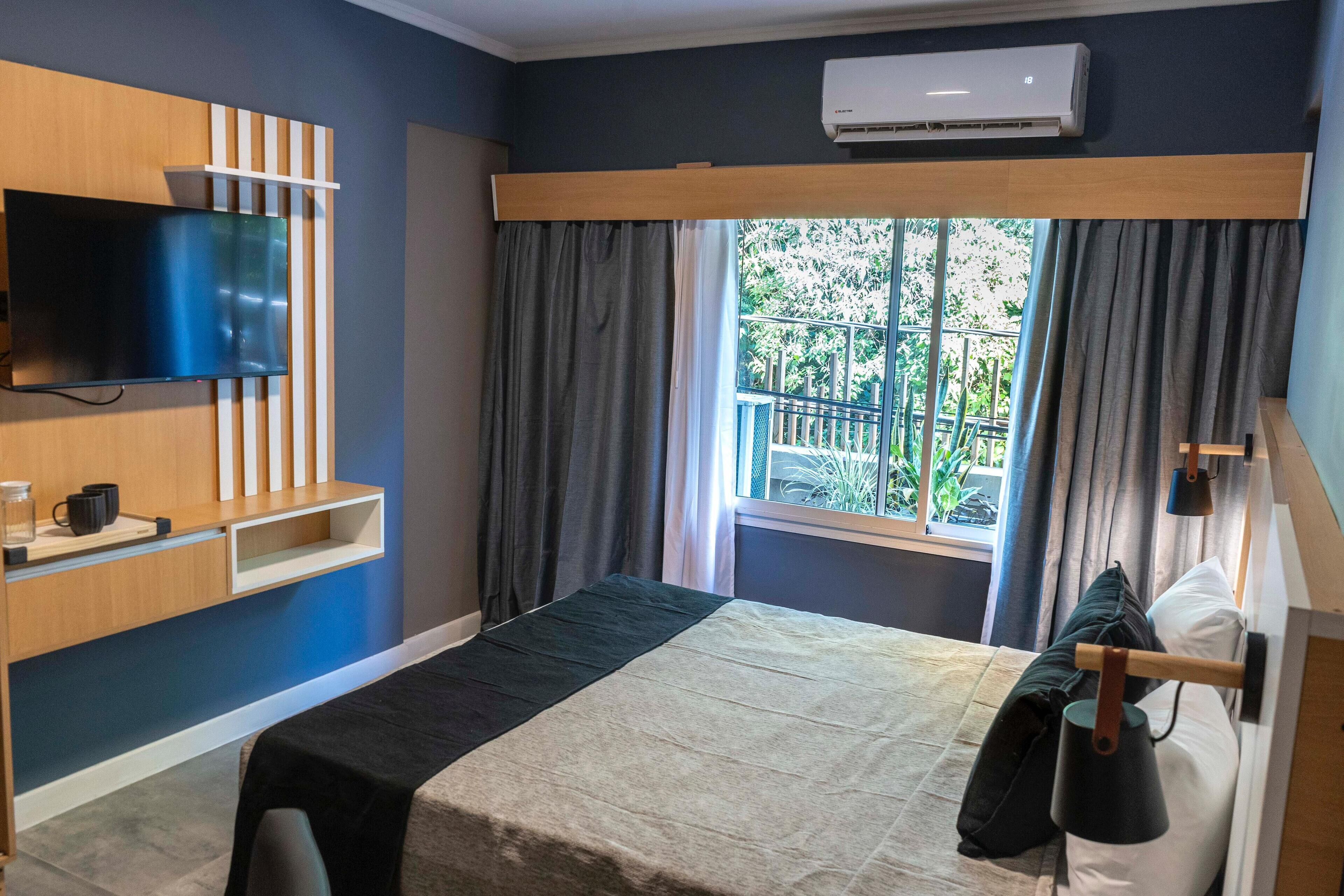 A hotel room with a bed, a TV mounted on the wall, a window with curtains, and an air conditioning unit.
