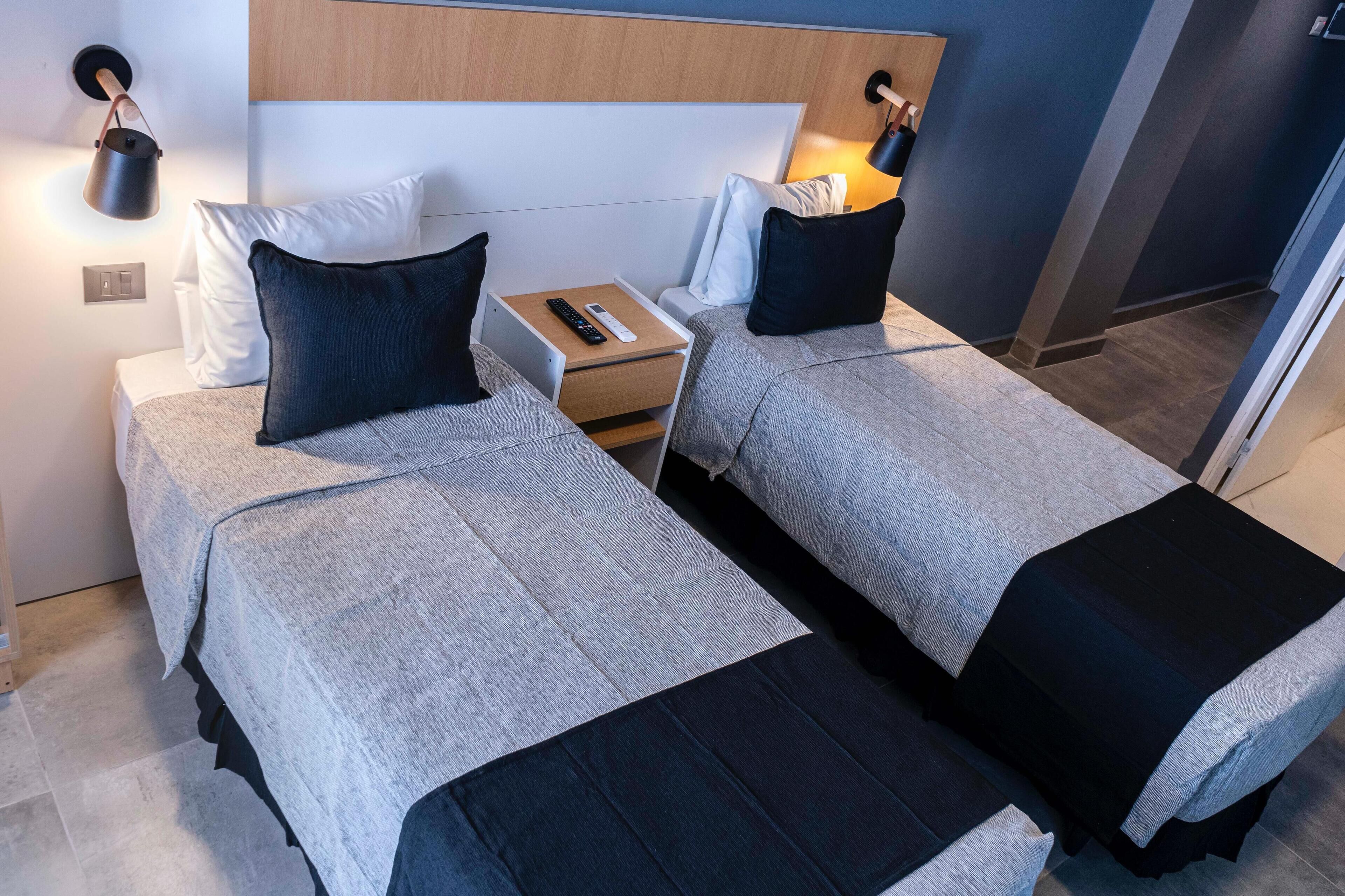 A hotel room with two beds, each with a pillow, a nightstand with a remote control, and a wall-mounted lamp.