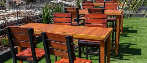 A rooftop terrace with wooden tables and chairs, surrounded by green artificial grass, and overlooking a cityscape.