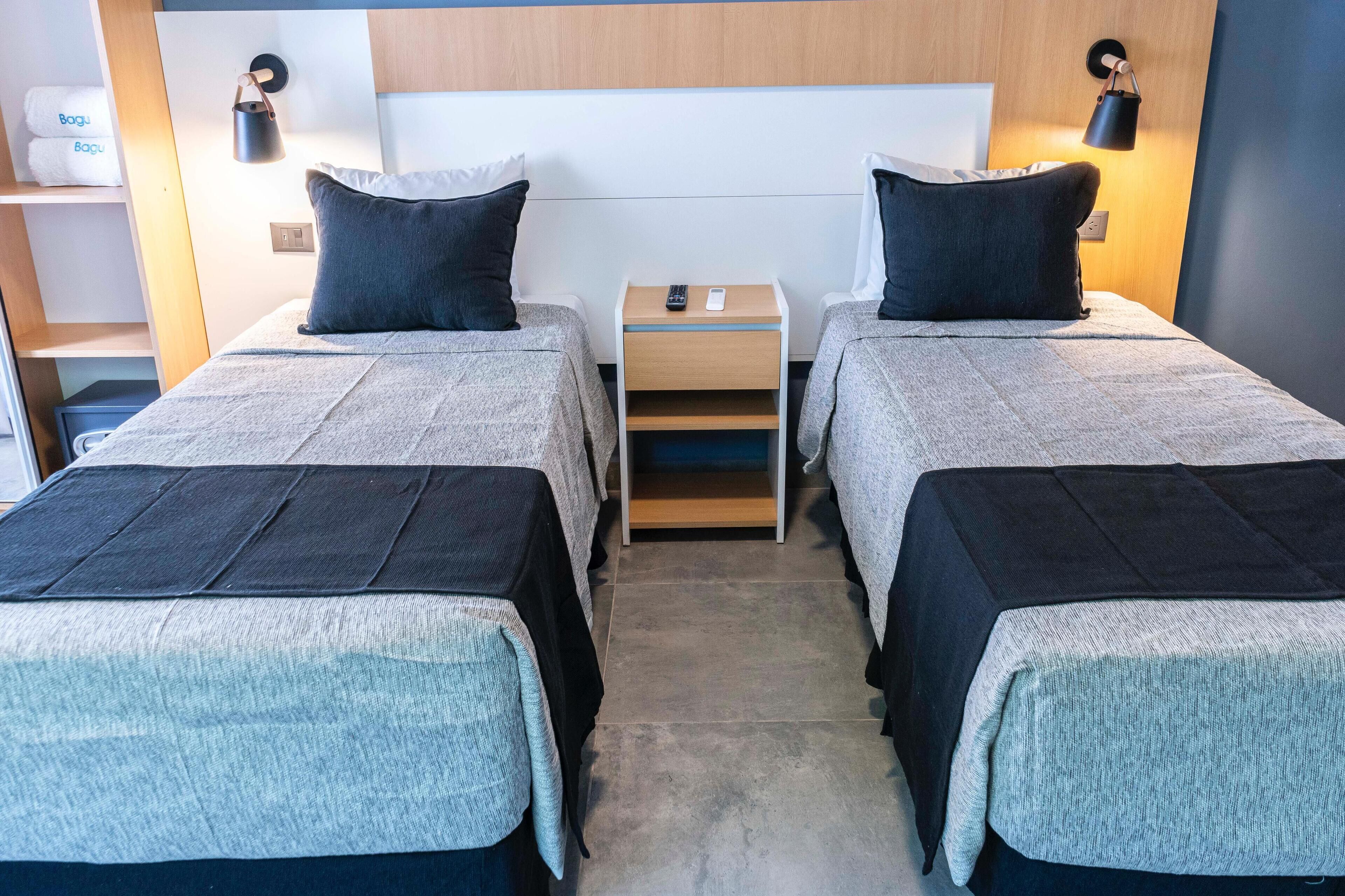 A hotel room with two beds, each with a pillow and a blanket. There is a nightstand with a remote control and a lamp between the beds.