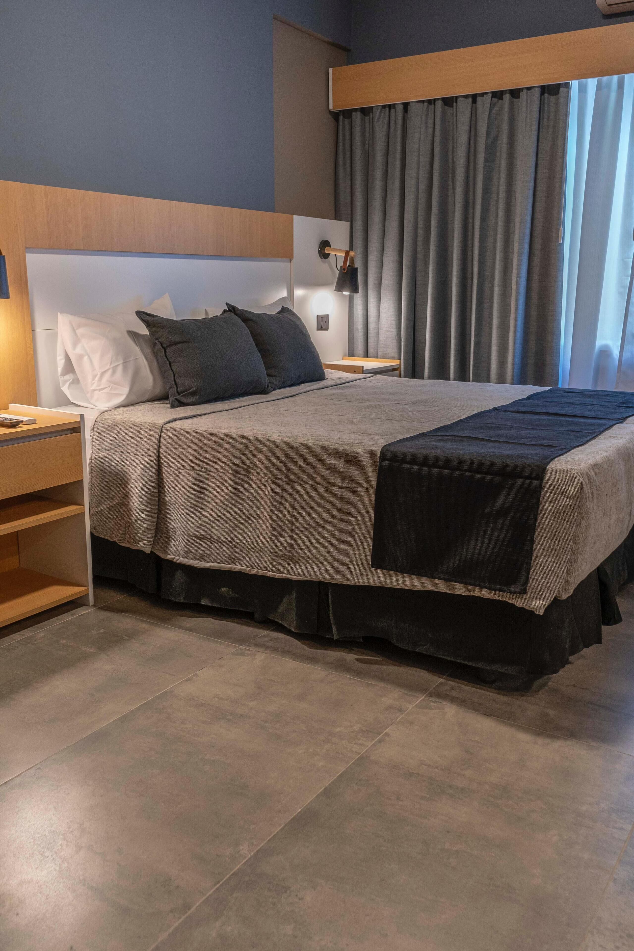 Un letto rifatto con cura, con copriletto grigio e nero, una testiera in legno e un comodino con una lampada.