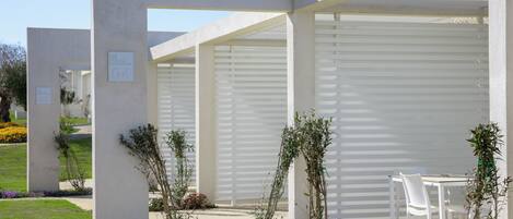 A modern building with white exterior walls and large garage doors. There are small plants and a pathway leading to a seating area with white chairs.