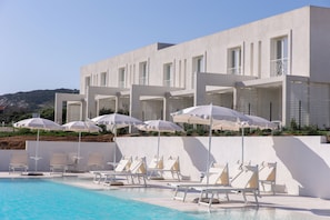 A modern hotel with a pool area featuring sun loungers and umbrellas, a white exterior, and a view of hills in the background.