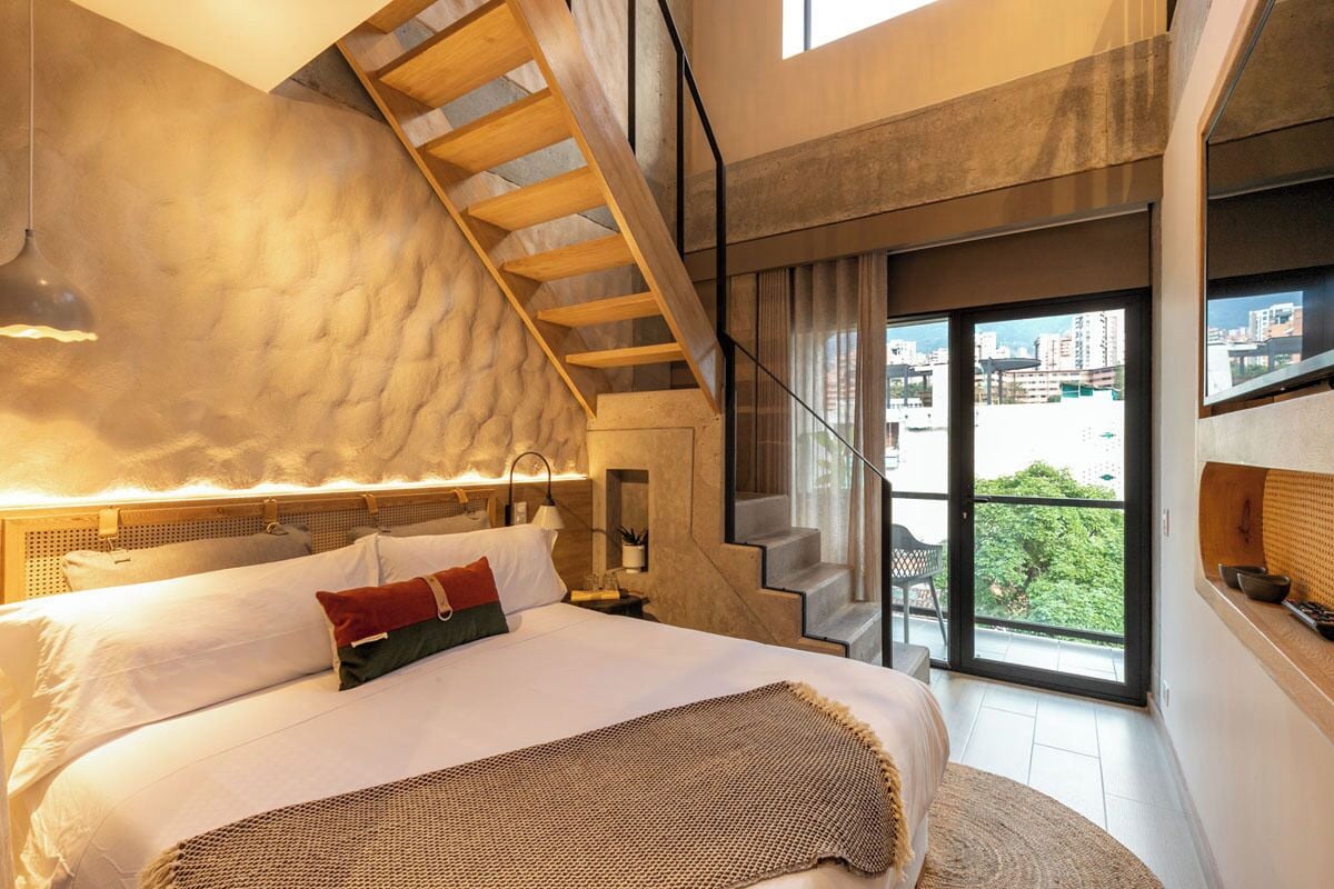A modern bedroom with a large bed, a wooden staircase, and a city view through the window.