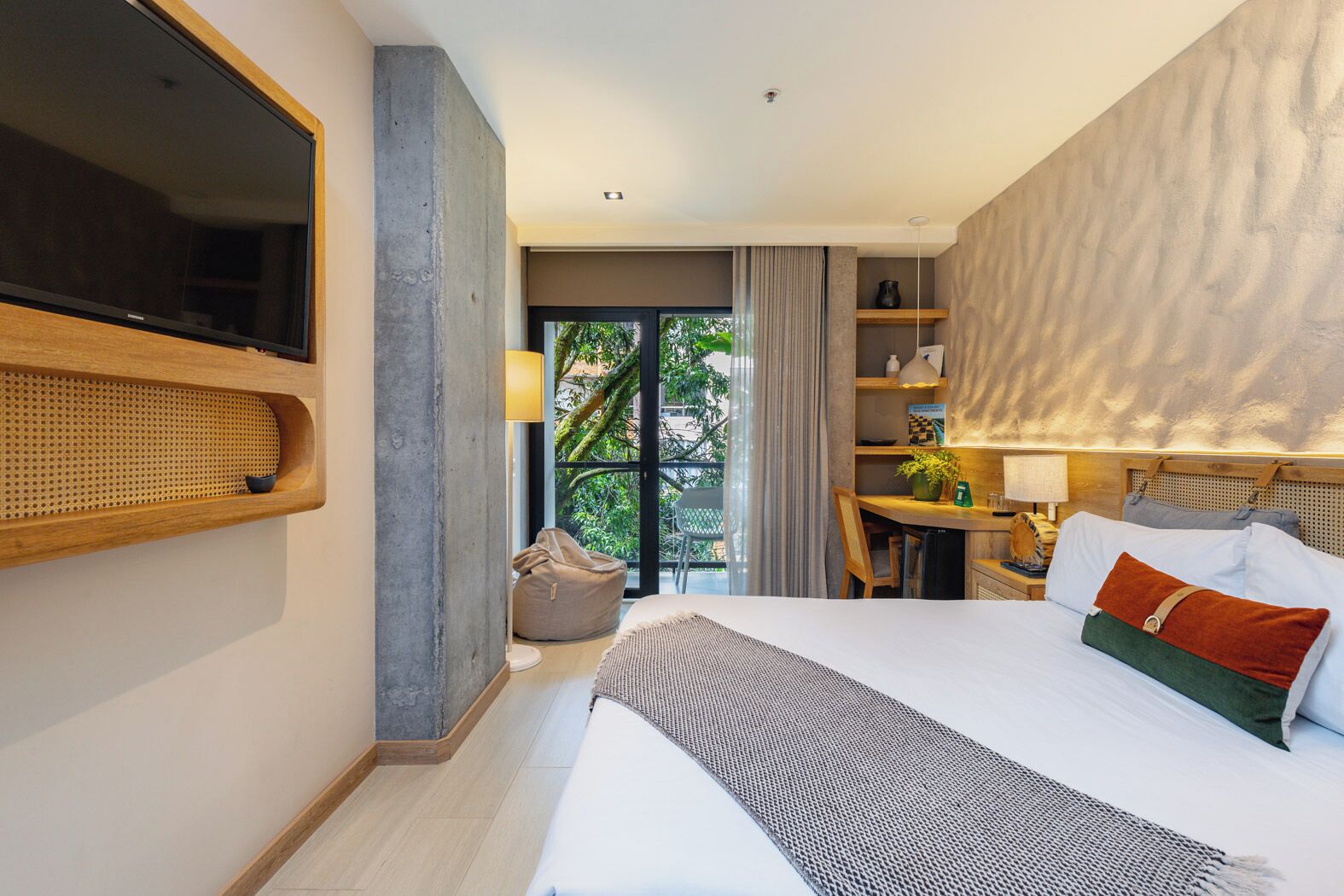 Quarto de hotel moderno com uma cama grande, televisão fixada na parede, escrivaninha com cadeira e vista para o verde lá fora.