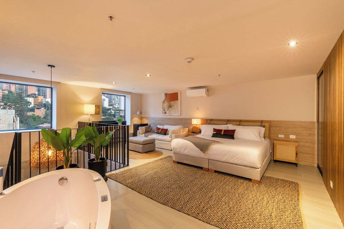 A modern hotel room with a large bed, a separate sofa, a small table, a bathtub, and a city view.