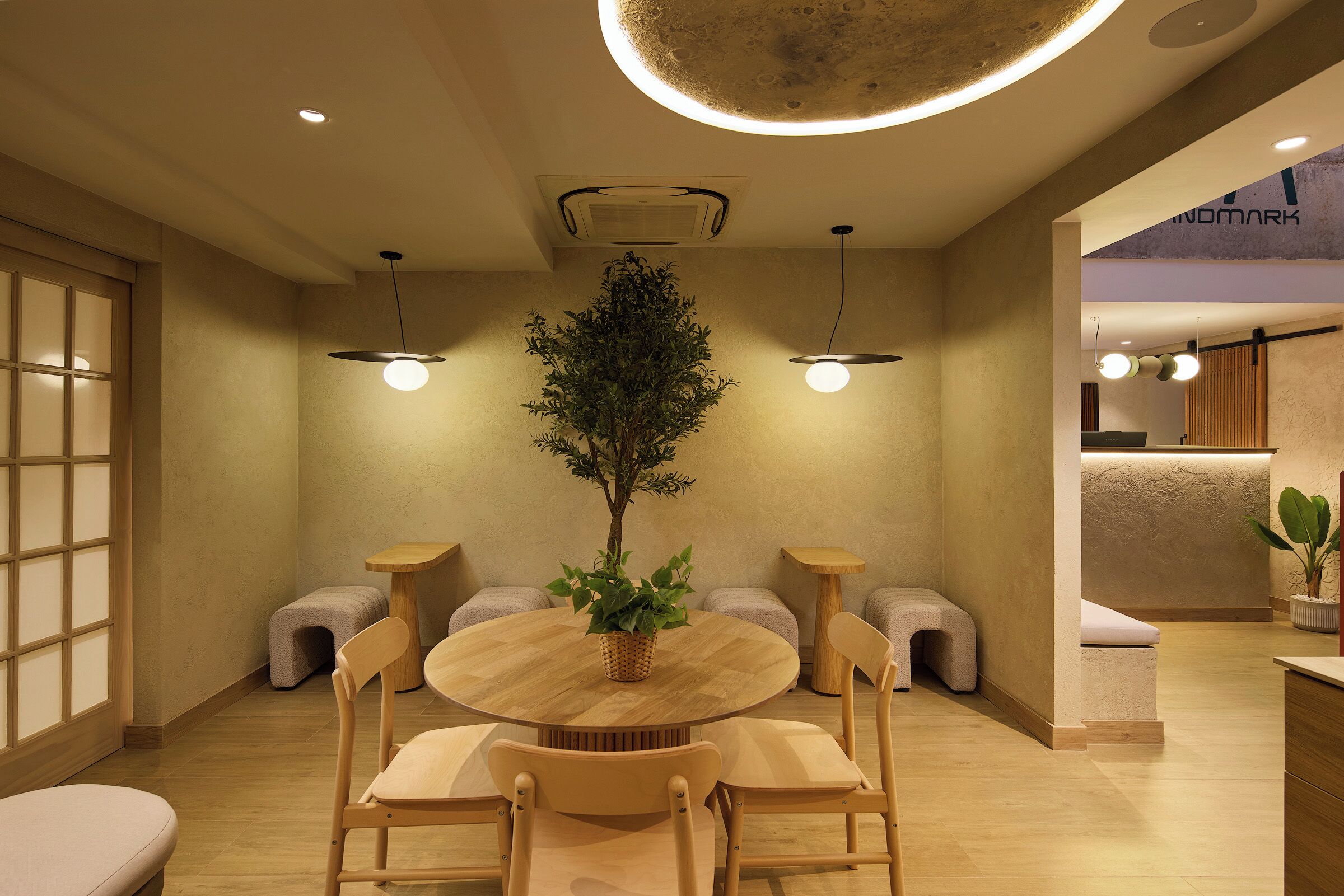 A modern interior with a round wooden table, cushioned chairs, a potted plant, recessed ceiling lighting, and a visible reception area.