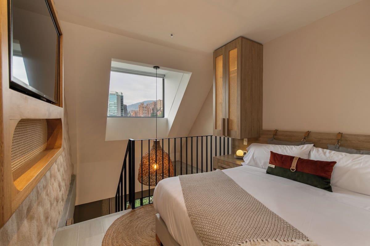 A modern hotel room with a large bed, a TV, a window with a city view, a wooden cabinet, and a unique hanging light fixture.