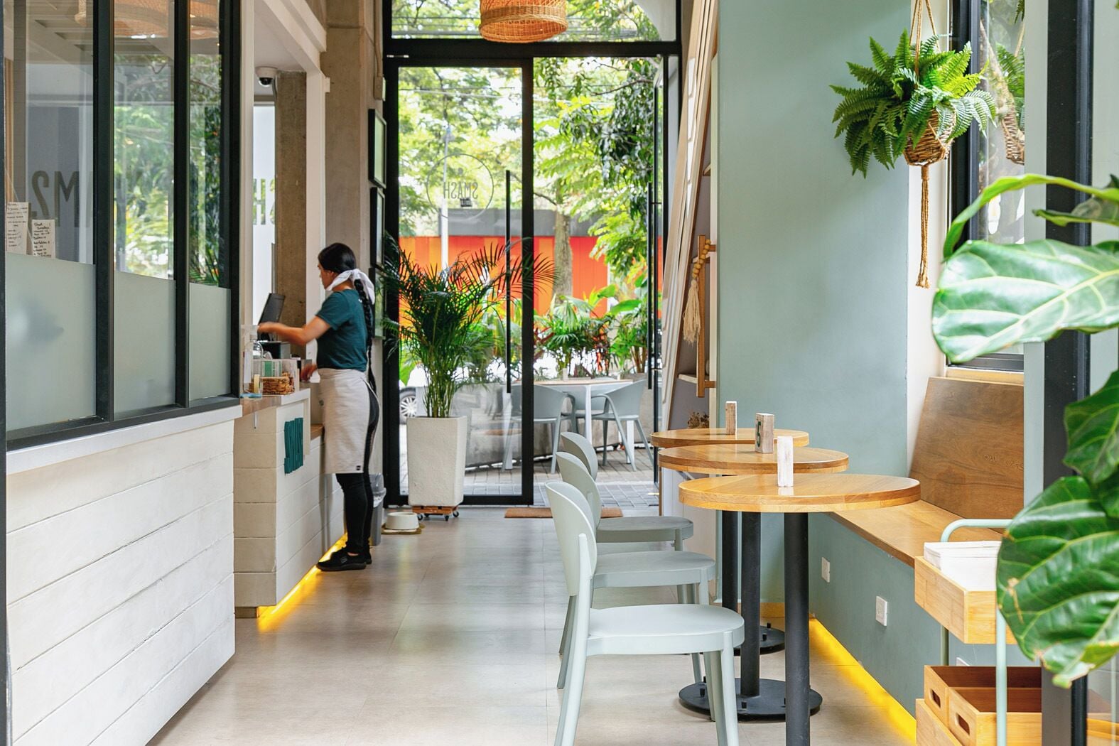 A modern cafe interior with a barista preparing drinks, white chairs, wooden tables, and potted plants.