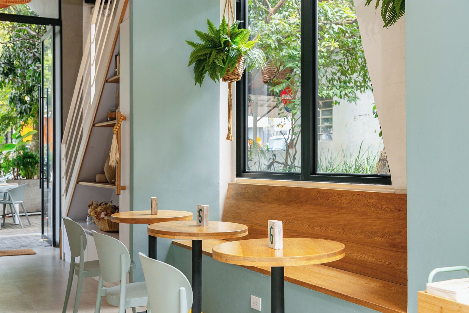 A cozy cafe with round wooden tables and white chairs, a large window with a view of greenery, and a hanging plant.
