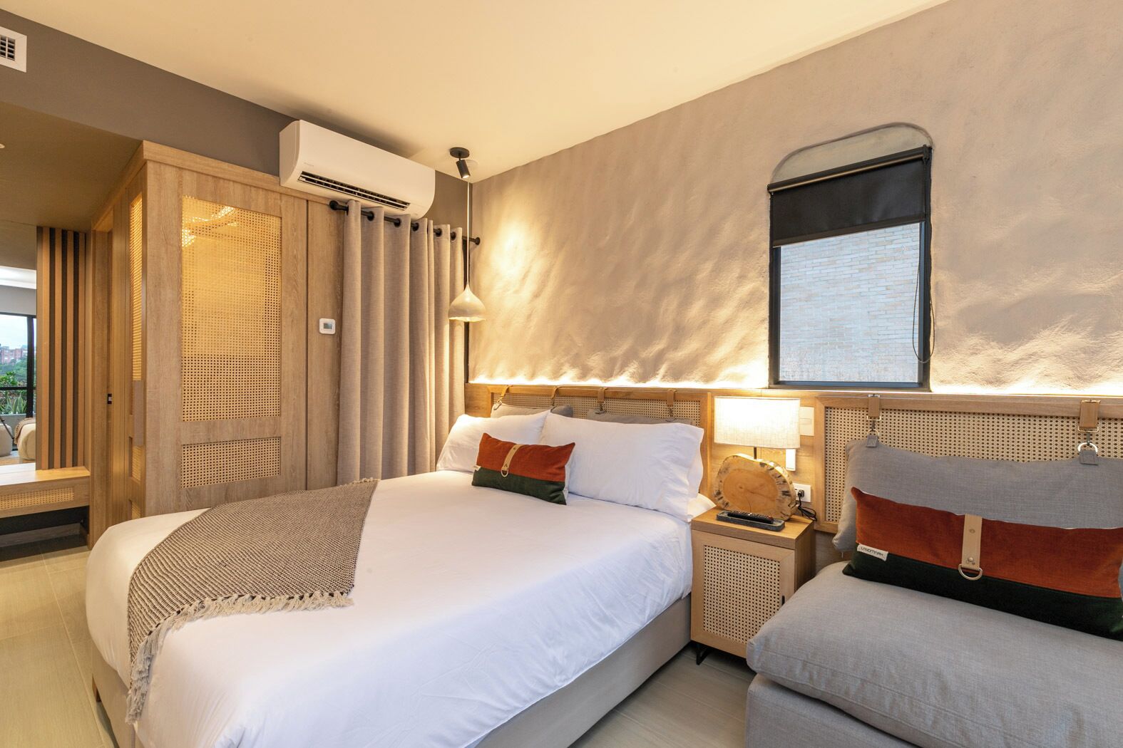 A hotel room with a large bed, a bedside table with a lamp, a wall-mounted air conditioner, and a window with a view.