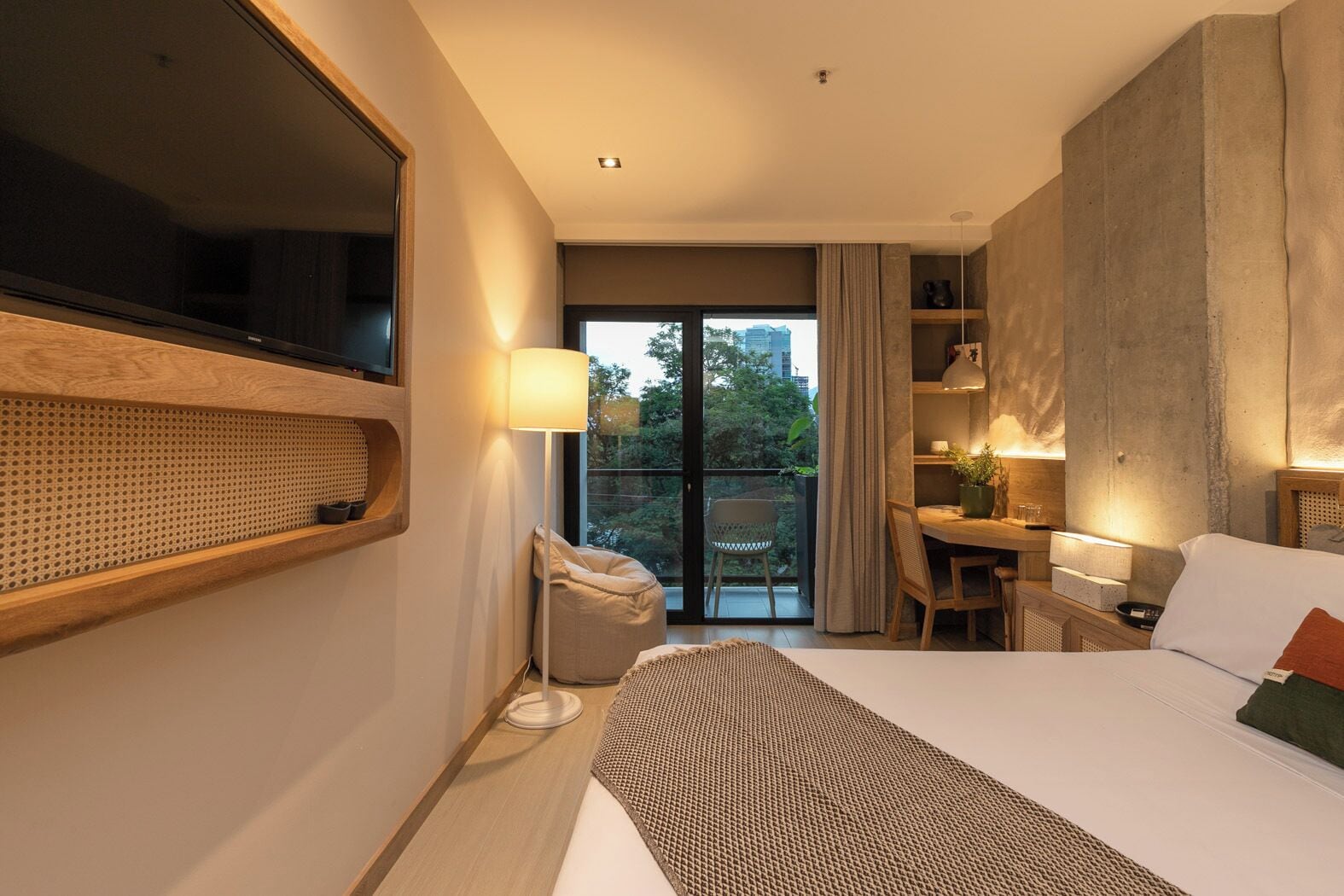 Quarto de hotel moderno com uma cama grande, uma escrivaninha, uma cadeira e varanda com vista para a cidade.