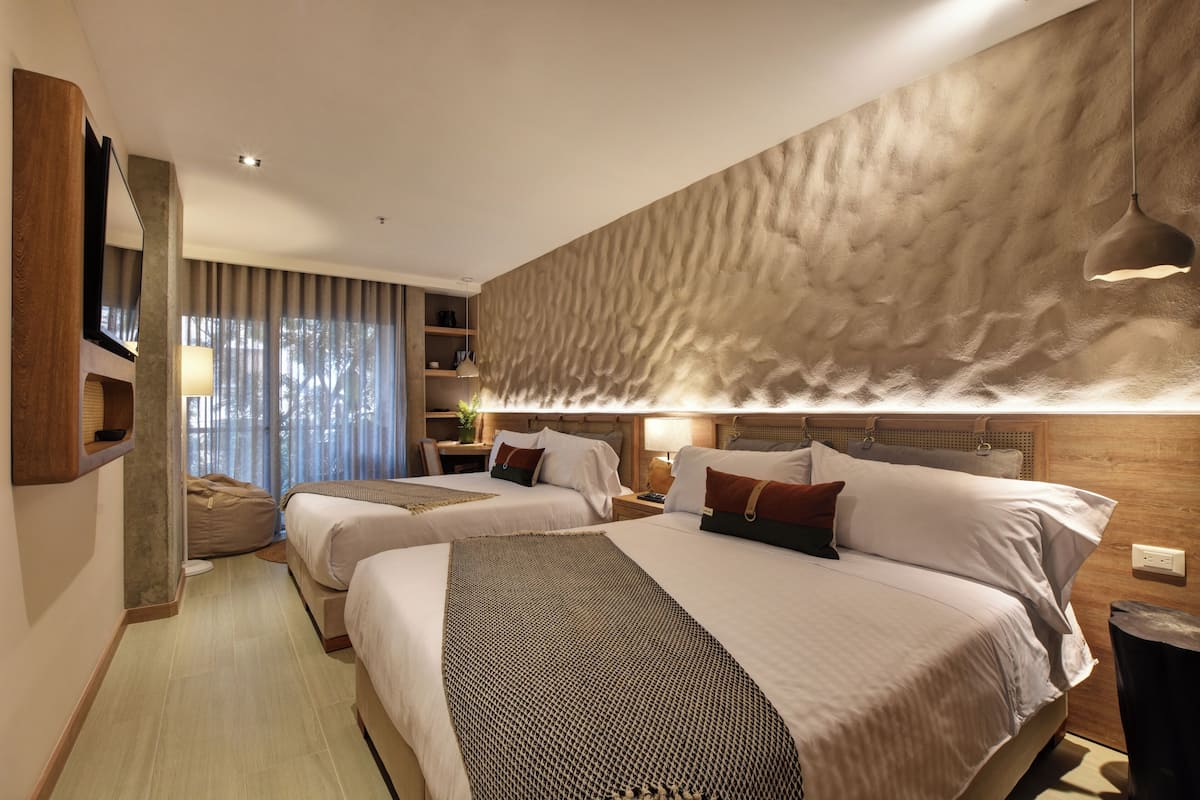 A hotel room with two beds, a wooden headboard, a large window with curtains, a bedside table with a lamp, and a wall-mounted TV.