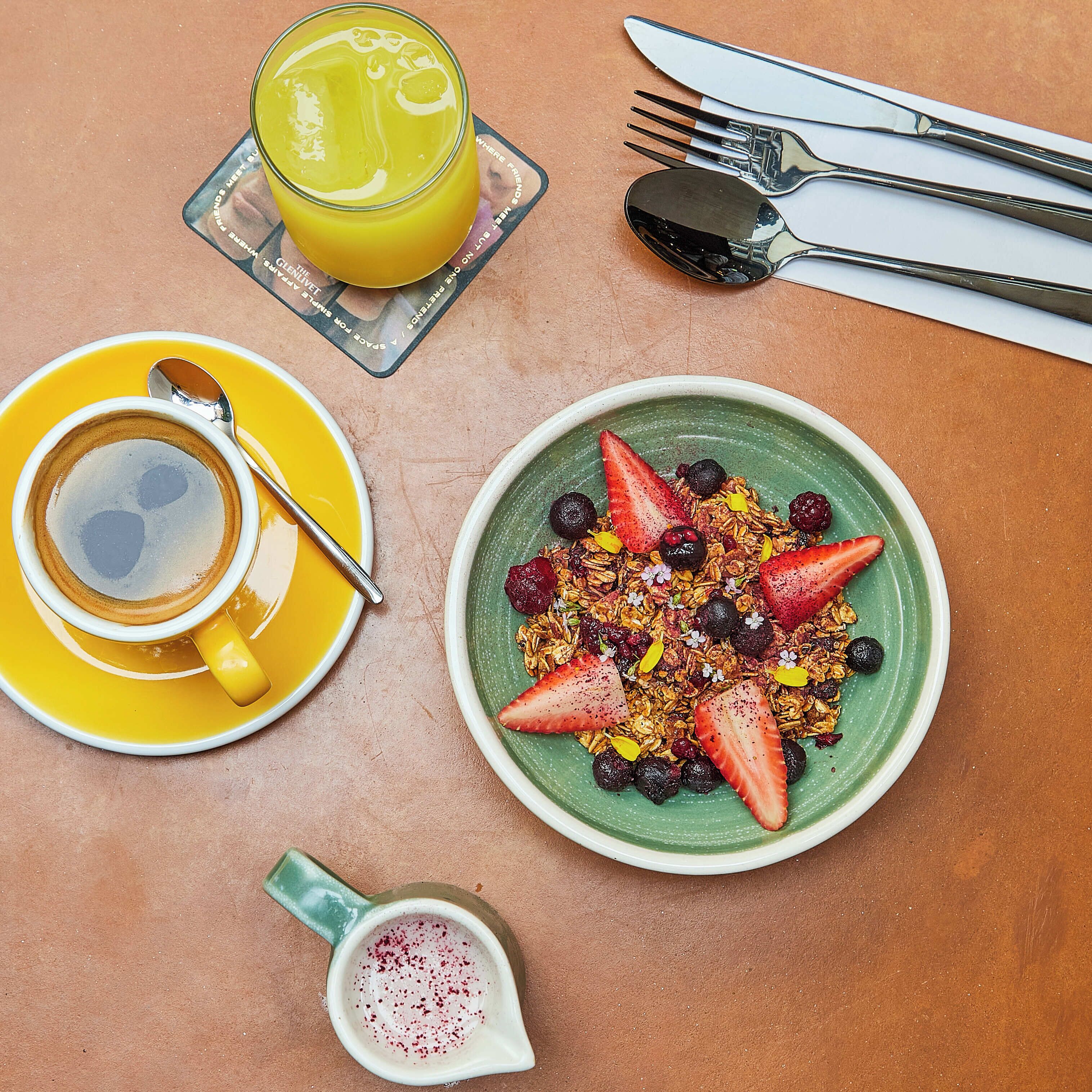 A breakfast setting with a cup of coffee, a glass of orange juice, a bowl of granola with berries, and a pitcher of a red beverage.