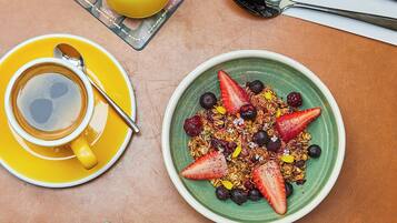 Un desayuno con una taza de café, un vaso de zumo de naranja, un bol de granola con bayas y una jarra de una bebida roja.