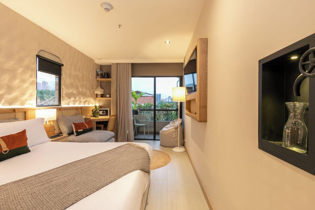 A modern hotel room with a large bed, a desk, a balcony with a view, and a mounted TV.
