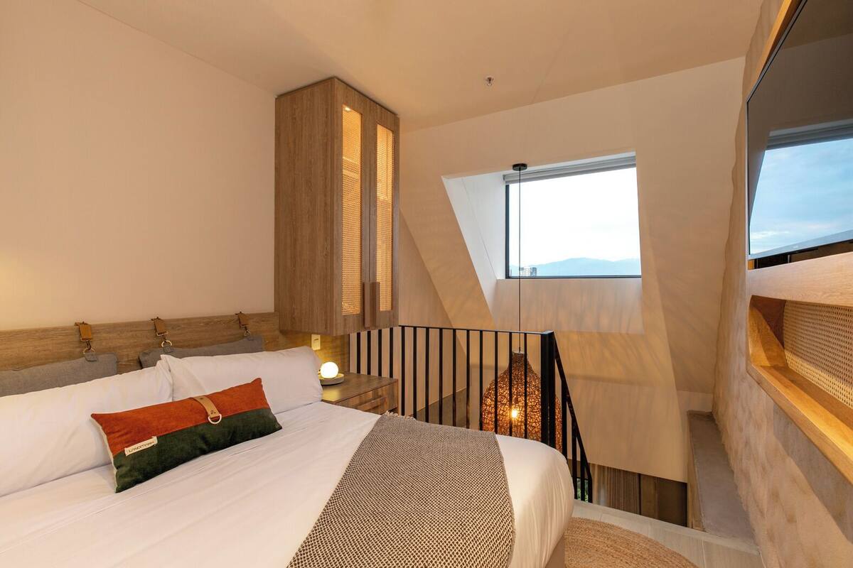 A bedroom with a large bed, a wooden headboard, a nightstand, and a window with a view of the sky.