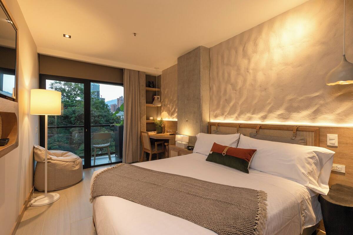 A modern hotel room with a large bed, a desk, and a balcony with a view of the city.