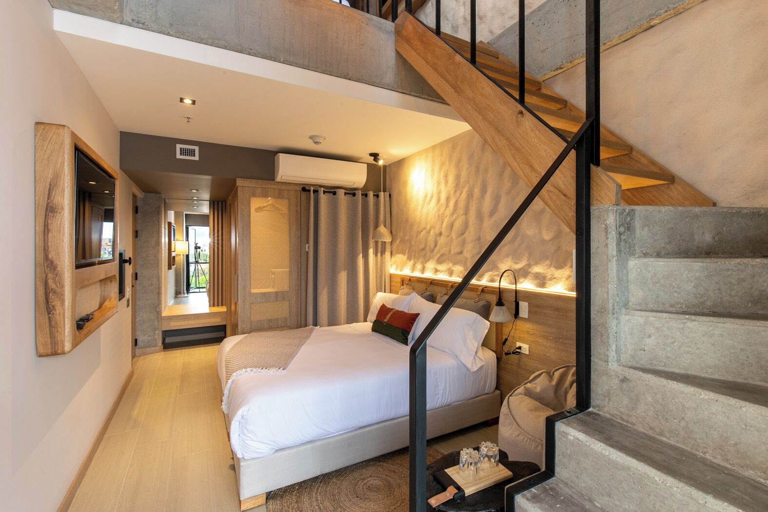 A modern bedroom with a large bed, a wooden staircase, and a view of the outdoors.