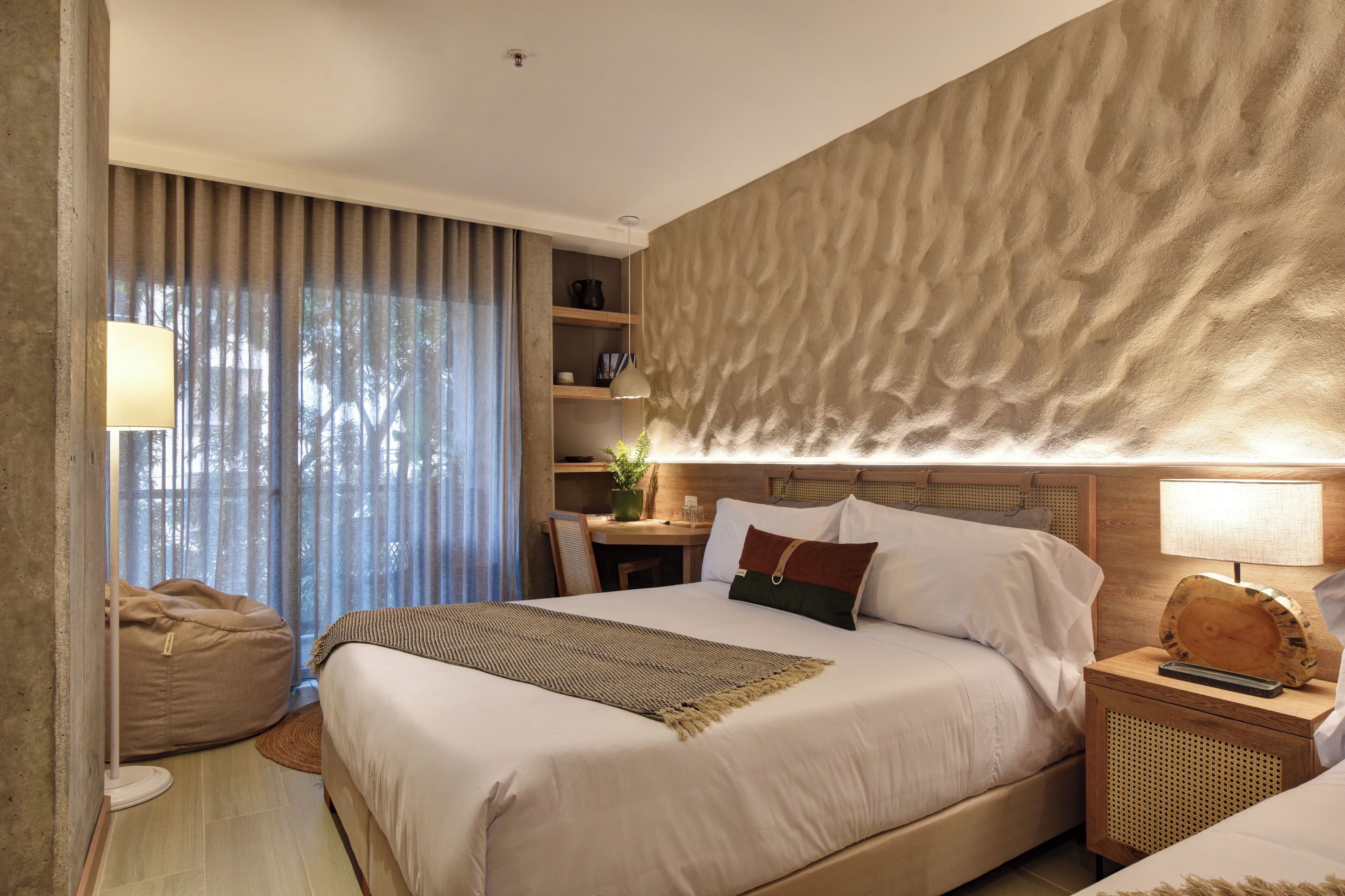 A hotel room with a large bed, a desk, a chair, a bean bag, and a wall with a textured pattern.
