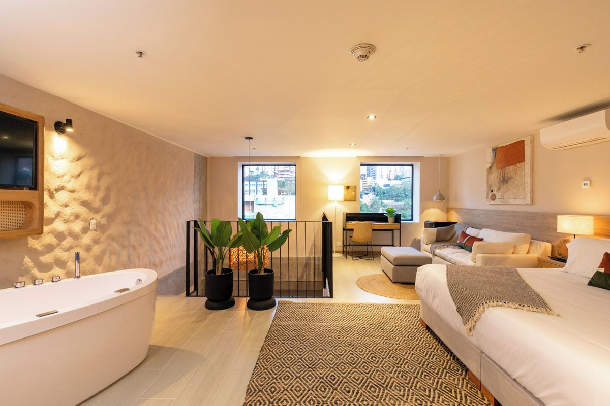 A modern hotel room with a large bathtub, a desk with a chair, a sofa, and a bed. There are potted plants and a patterned rug on the floor.