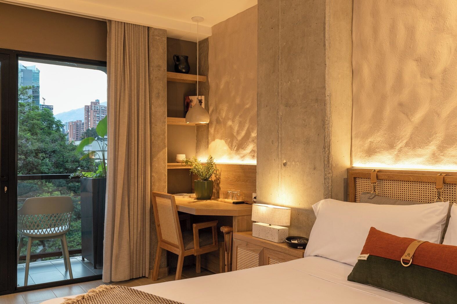 A hotel room with a balcony, a desk, a bed, and a view of the city.