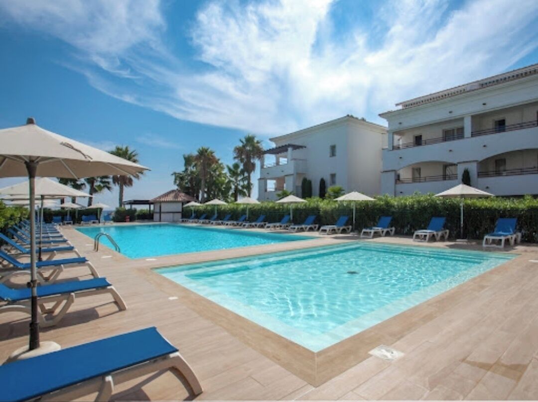 Un área de piscina con sillones de playa y sombrillas, cielo despejado y un edificio de varios pisos al fondo.