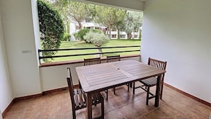 Un balcón con mesa y sillas de madera, vista a un césped verde y edificios a lo lejos.